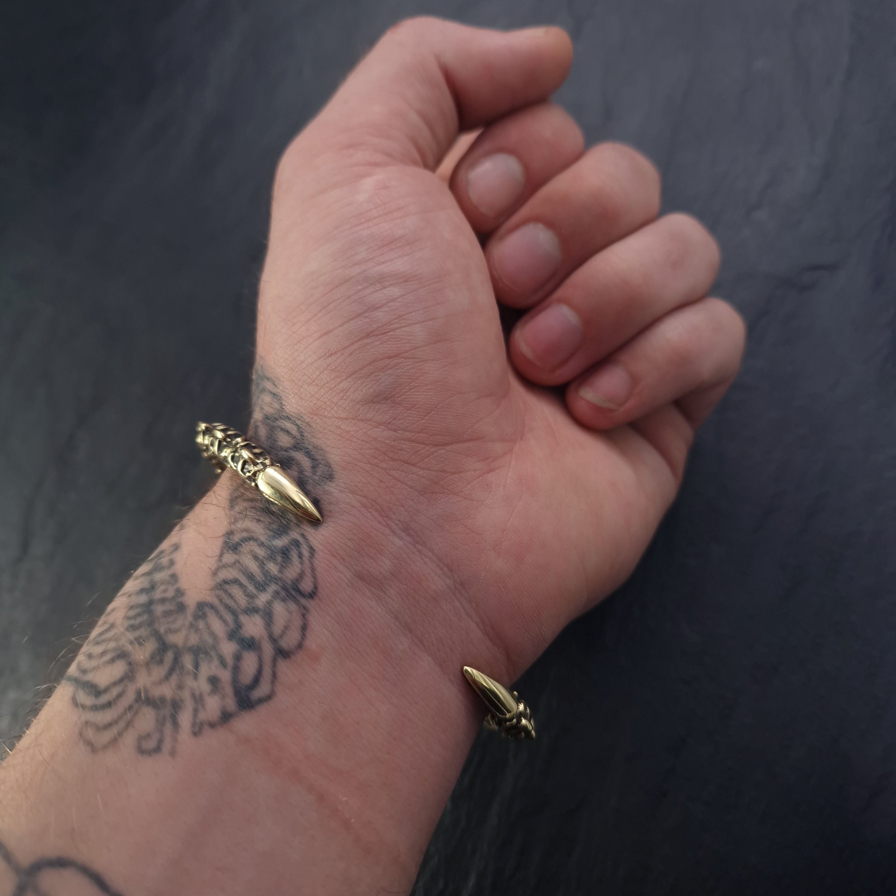 A hand with a bird tattoo on the wrist is shown, wearing the SHIRO Men's Gold Cuff Bracelet by Kaya Movement, which features pointed ends and a sleek design reminiscent of a reptile spine. The background is a dark, smooth surface.
